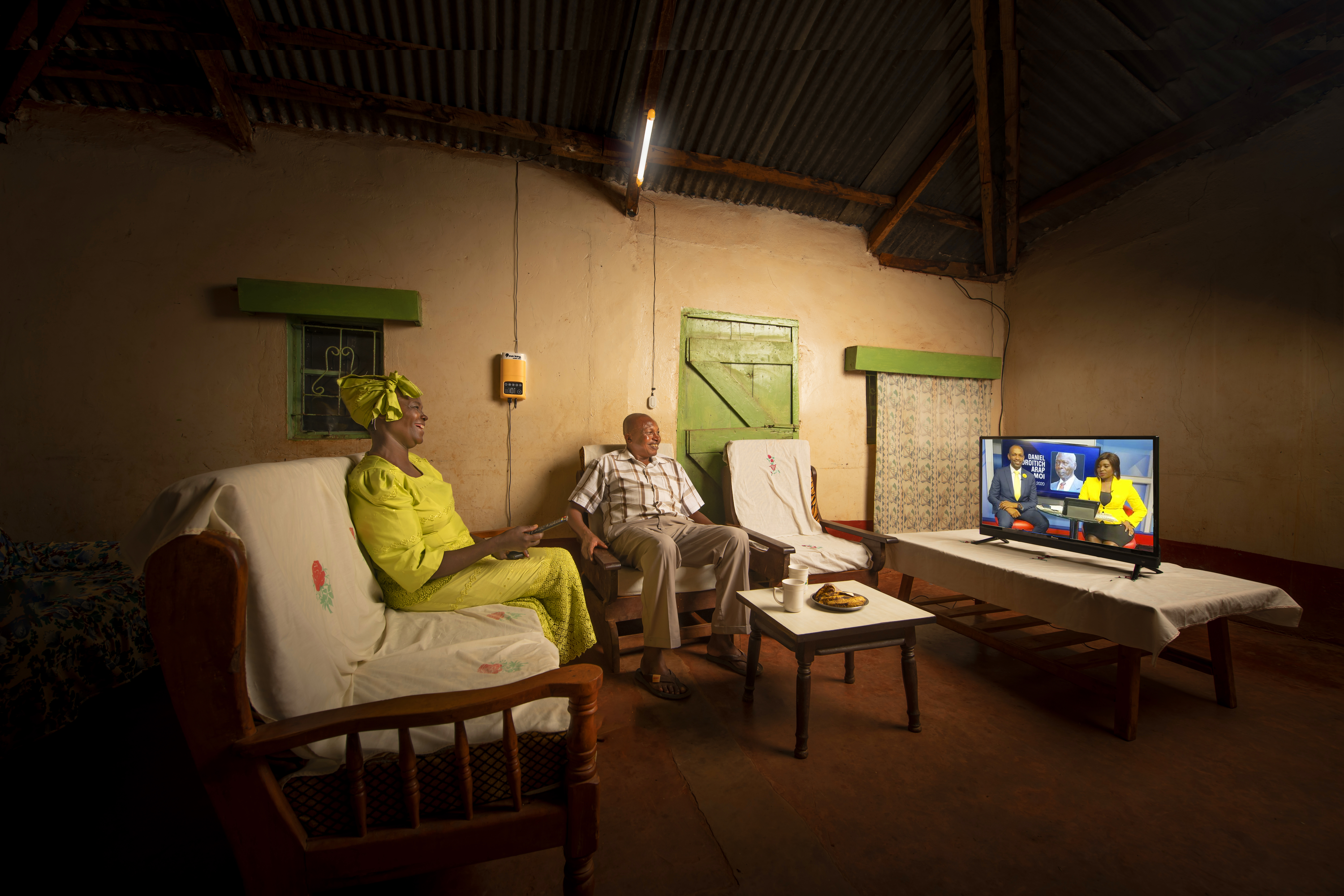 A couple sit in their living room watching the TV, with the Sun King 500X solar home lighting system and HD TV.